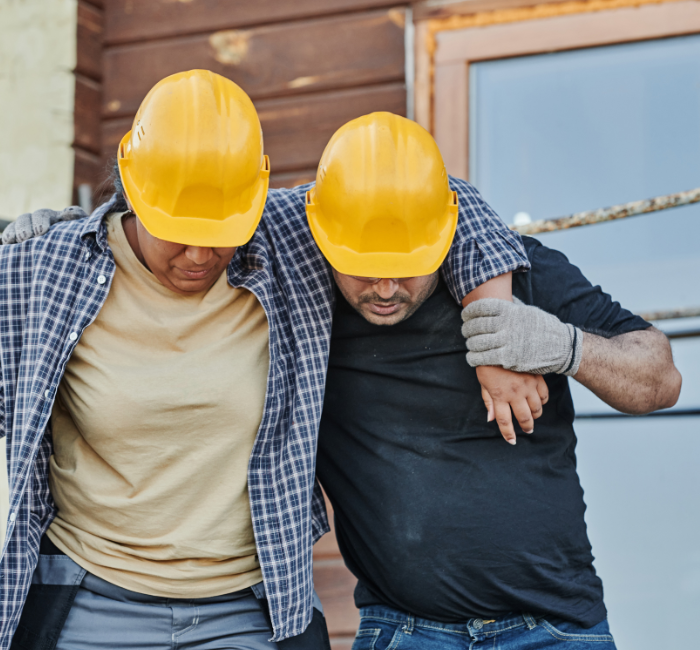 Injured worker on building site wearing a yellow hard-hat being helped by colleague